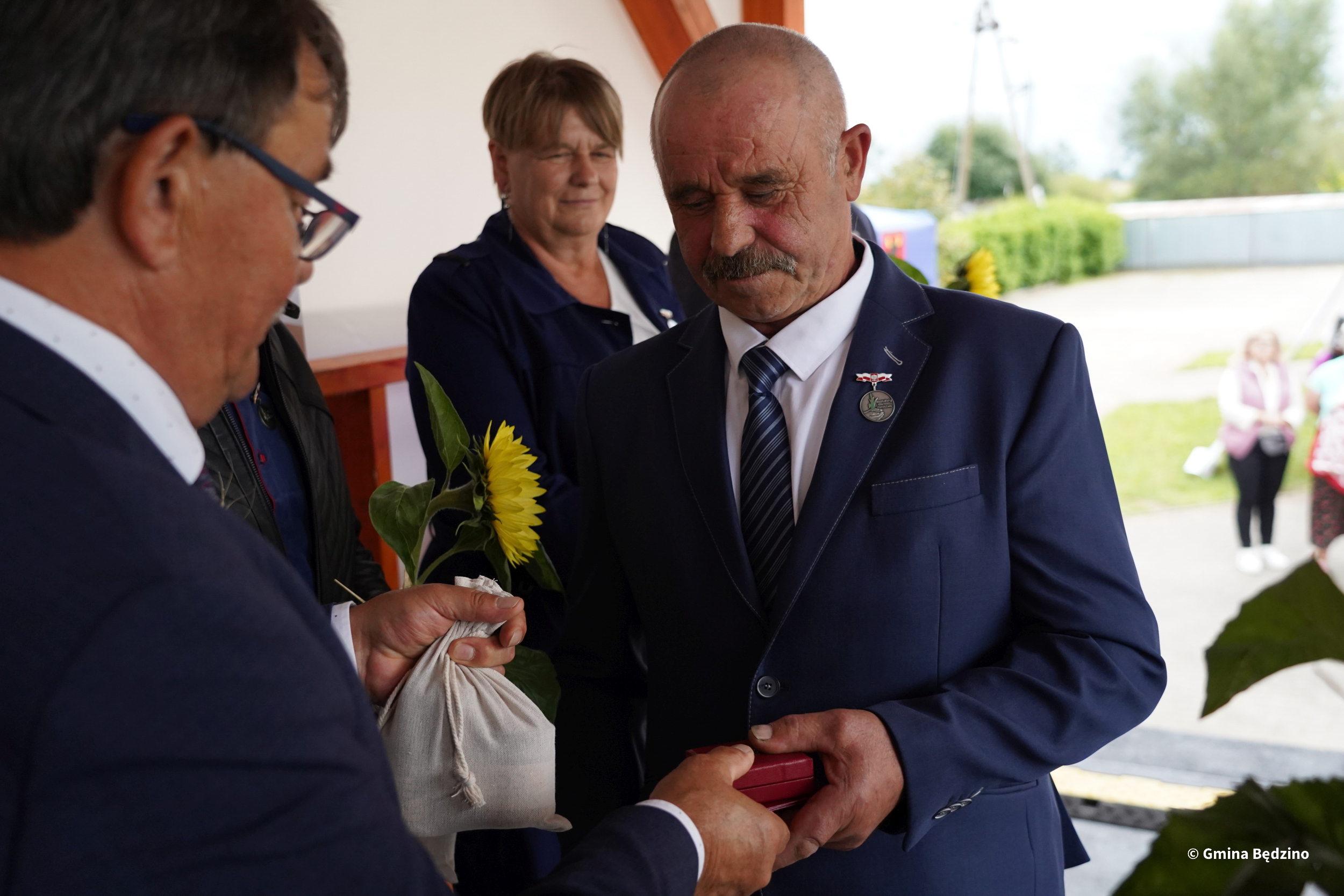 Mężczyzna odbiera nagrodę na Dożynkach Gminnych Tymień 2025. Wręczono mu małe pudełko i słonecznik.