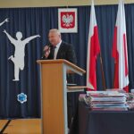 Mężczyzna przemawiający na podium podczas uroczystości zakończenia roku szkolnego w Będzinie. Widoczne są polskie flagi.