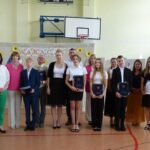 Uczniowie i nauczyciele na uroczystości zakończenia roku szkolnego w szkole w Będzinie. Widoczny baner z napisem „Wakacje”.