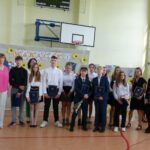 Uczniowie i nauczyciele na uroczystości zakończenia roku szkolnego w szkole w Będzinie. Widoczny baner z napisem "Wakacje!".