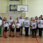 Uczniowie i nauczyciele na uroczystości zakończenia roku szkolnego w szkole w Będzinie. Widoczny baner z napisem "Wakacje!".