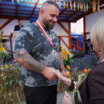 Mężczyzna odbiera nagrodę na Dożynkach Gminnych Tymień 2025. Uścisnął dłoń kobiety wręczającej medal.