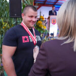 Mężczyzna odbiera nagrodę w Dożynkach Gminnych Tymień. Uśmiecha się, gdy kobieta wręcza mu medal.