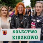 Trzy kobiety w gminie Dożynki Tymień trzymające tabliczkę z napisem „Sołectwo Kiszkowo”.
