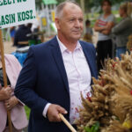 Mężczyzna w garniturze w Dożynkach Gminne Tymień, trzymający bukiet dożynkowy. Widoczny napis „Sołectwo Lasin Kosz”.