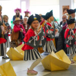 Dzieci przebrane za piratów występują na imprezie, trzymają pirackie flagi i stoją obok papierowych łódek.
