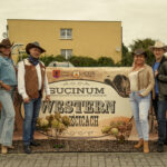 Grupa osób ubranych w stroje kowbojskie pozująca przed szyldem z napisem „Western”.